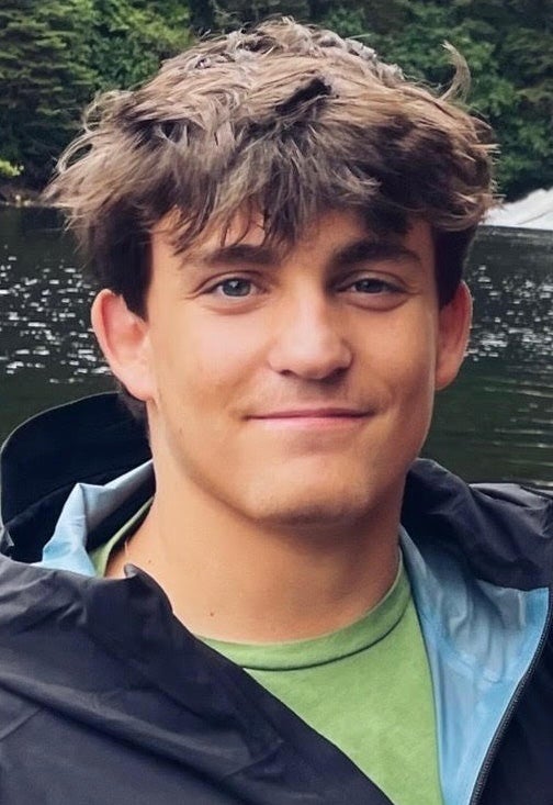 A young man with dark wavy hair, wearing a green t-shirt under a blue rain jacket.