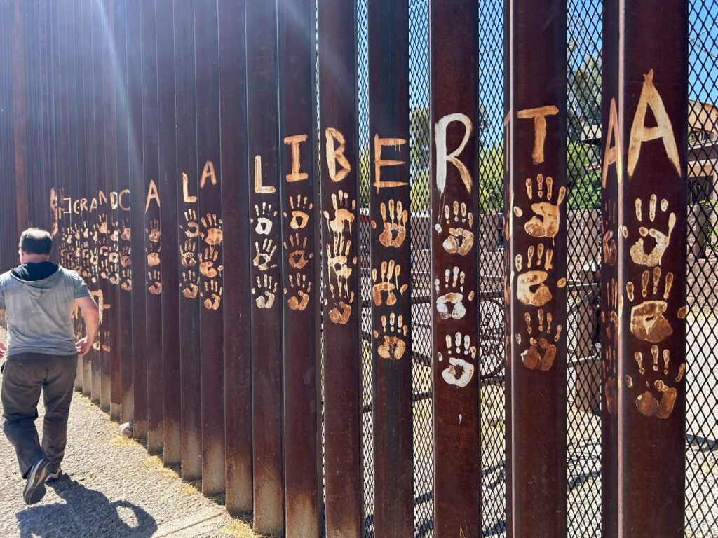 A man walks along the Mexico-US border fence. He's dressed in grey-ish clothing. The fence is made of rusted steel and has graffiti written on it.