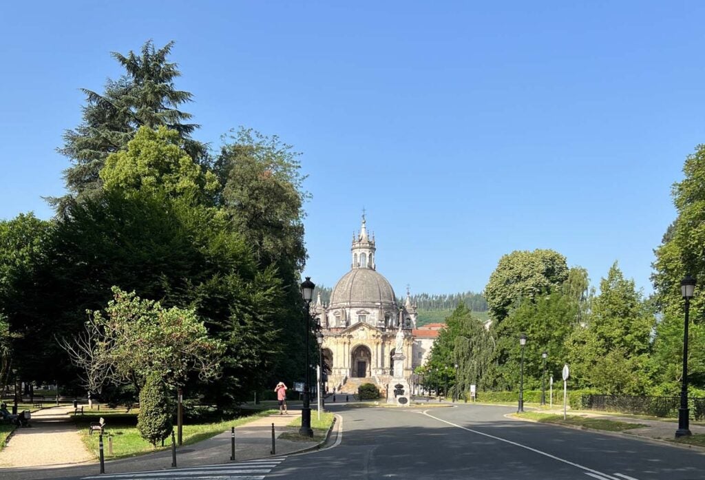 A tree-lined boulevard leading up to a Basilica with a grand staircase out front, a domed roof with a cross perched on top.